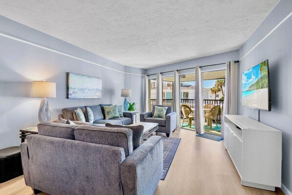 A modern living room with grey sofas, a flat-screen TV, and a view of the outdoor balcony.