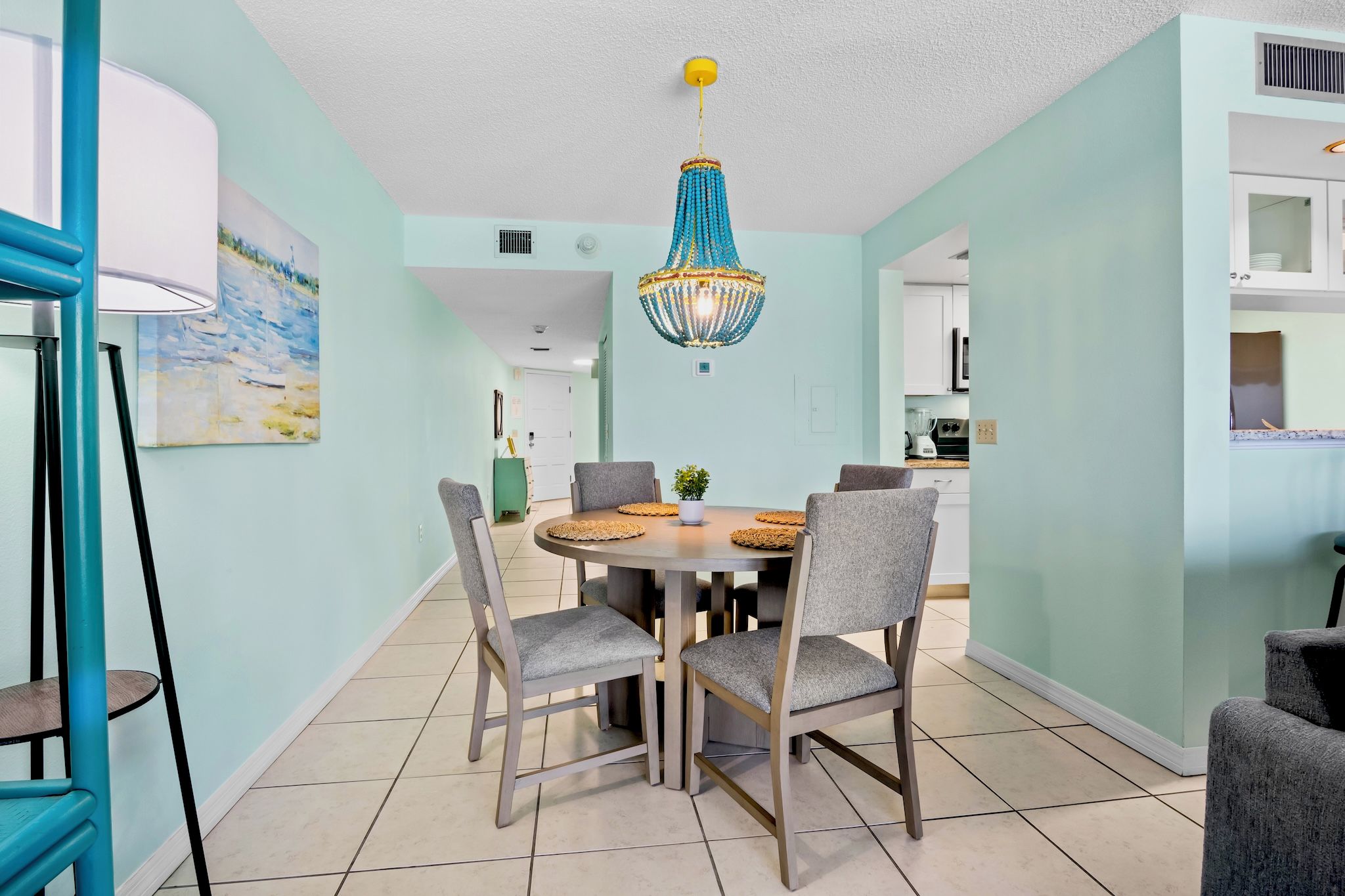 A dining area with a round table, four chairs, a chandelier, and a kitchen in the background.