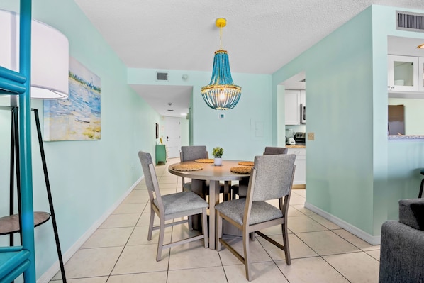 A dining area with a round table, four chairs, a chandelier, and a kitchen in the background.