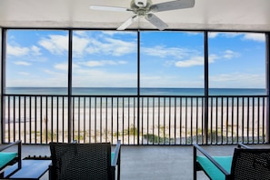 A room with a large window overlooking a beach, a ceiling fan, and two chairs.