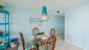 A dining area with a round glass table, four wooden chairs, a blue and green patterned tablecloth, a decorative blue and orange chandelier, and a wall-mounted painting of a beach scene.