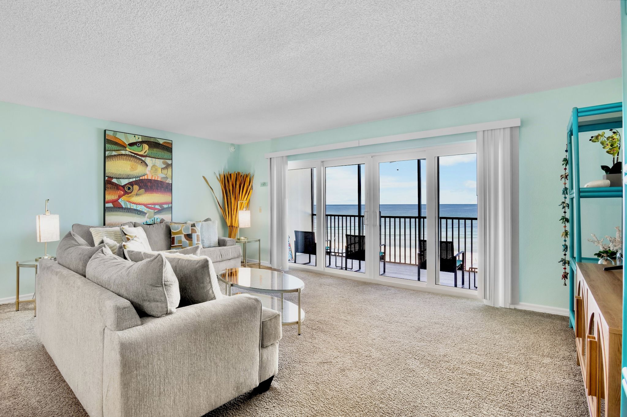 A living room with a sofa, a coffee table, and a painting on the wall. There is a balcony with a view of the ocean.