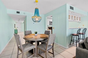 Un comedor con una mesa redonda, sillas tapizadas grises, una lámpara azul decorativa y una cocina al fondo.