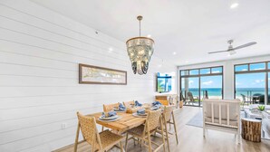 A dining area with a wooden table and chairs, a chandelier, and a view of the ocean through large windows.