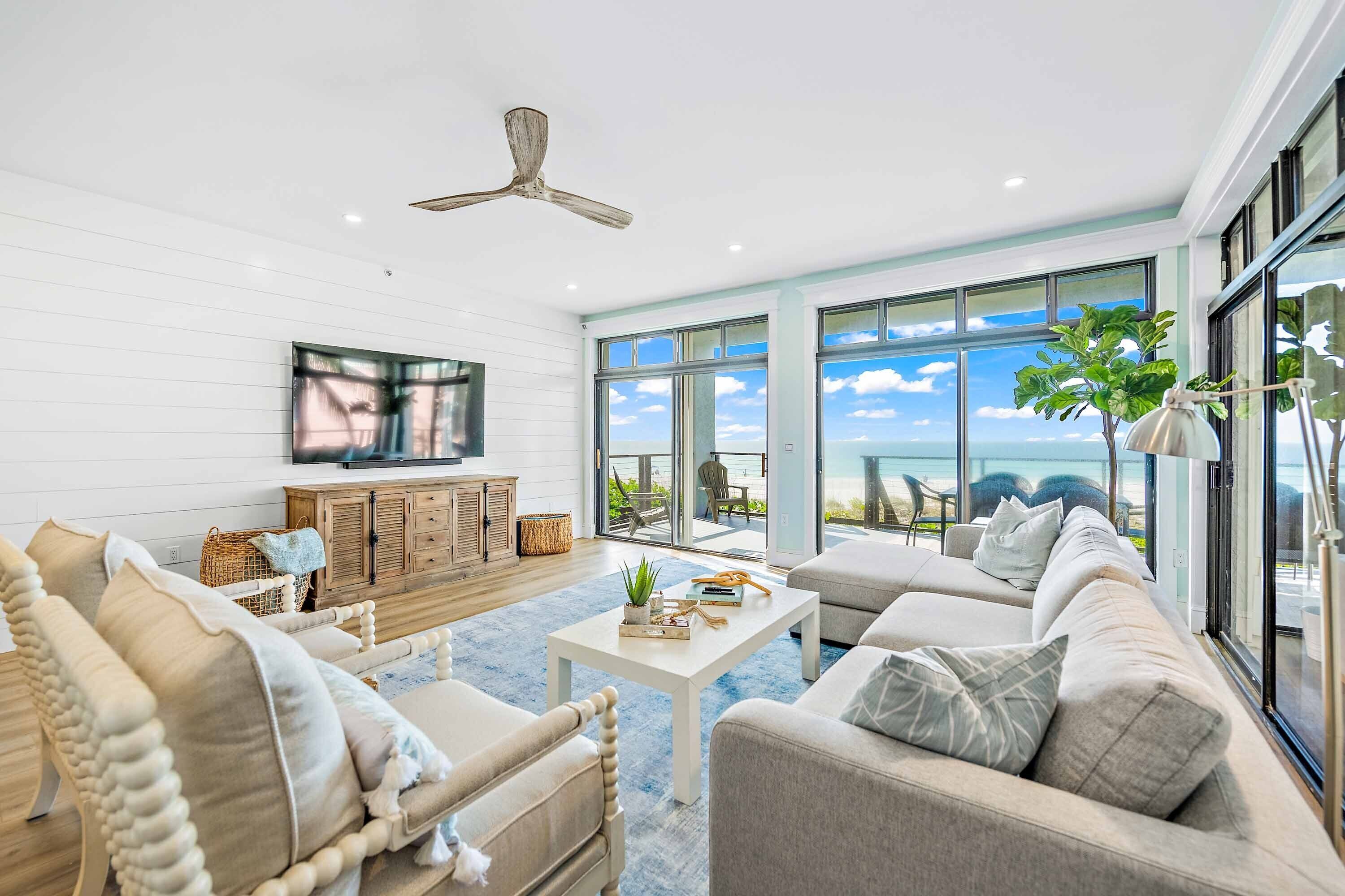 A spacious living room with a large sofa, a coffee table, and a television. There is a ceiling fan and large windows offering a view of the outdoors.