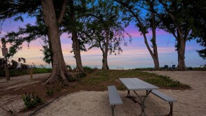 Una mesa de picnic con bancos en una zona arenosa rodeada de árboles, con vista al océano de fondo.