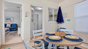 Un comedor con una mesa redonda, sillas y un paraguas azul. Se observa un sofá azul y un cuadro enmarcado al fondo.