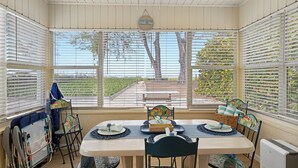 A sunroom with a table set for two, chairs with colorful patterns, a wicker basket, and a view of a garden through large windows.