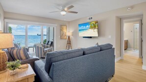Un salon avec un grand canapé gris, un téléviseur diffusant une scène de plage et une vue sur l’océan par de grandes fenêtres.