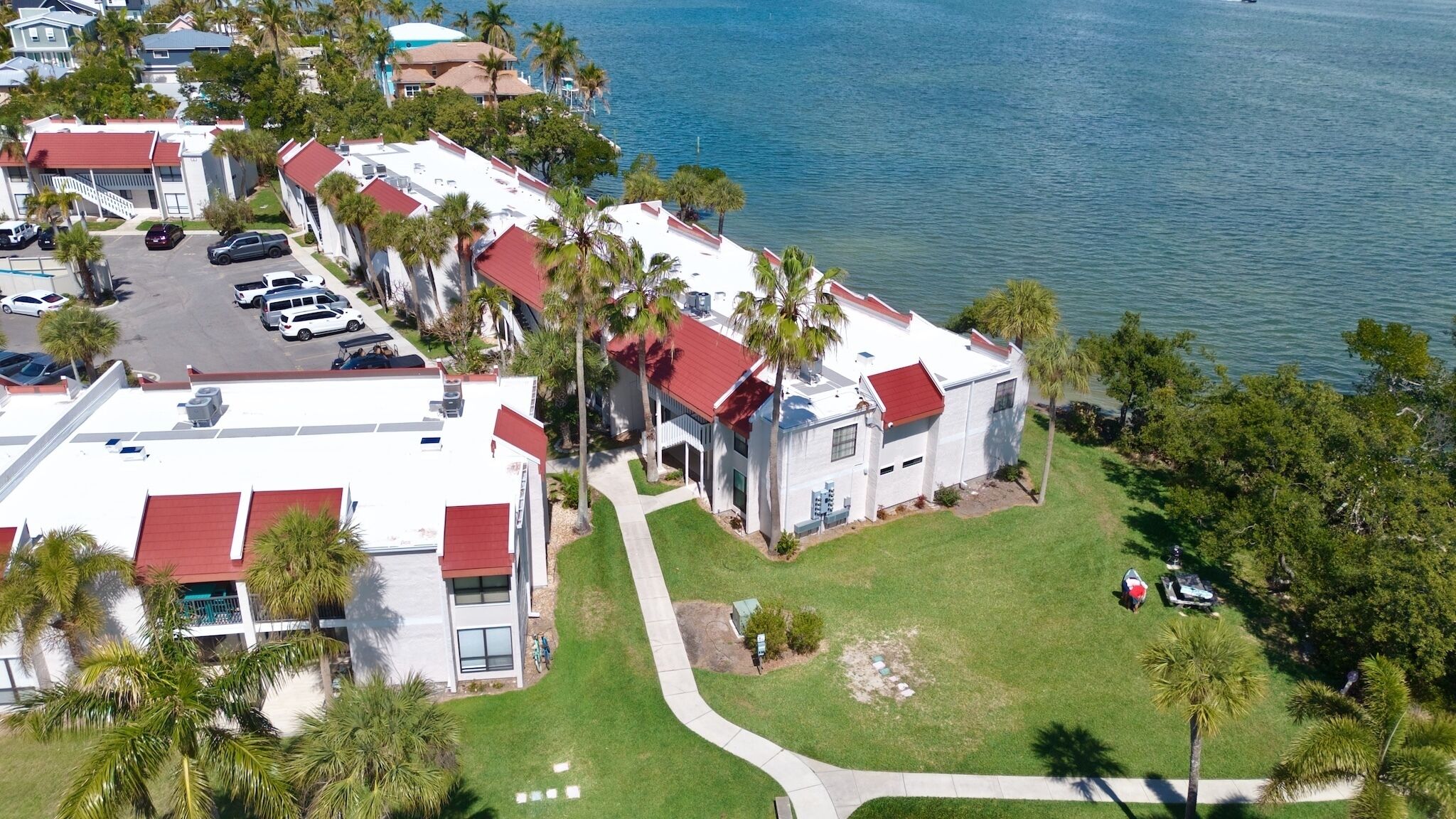 An aerial view of a coastal residential area with houses featuring red roofs, surrounded by palm trees and a body of water.