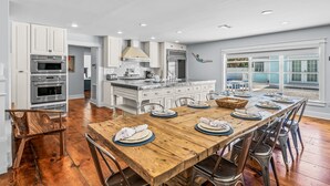 A spacious kitchen with white cabinetry, a wooden dining table set for six, and a view of the outdoors through large windows. - North End Home, 5 Houses to Beach! Near Pine Ave! (Anna Maria)