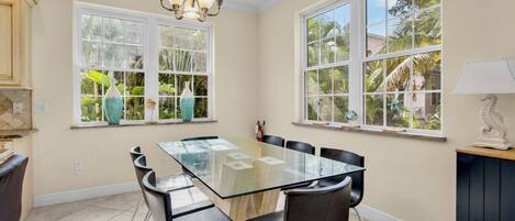 A dining area with a glass-top table, black chairs, a chandelier, and windows with tropical views.
