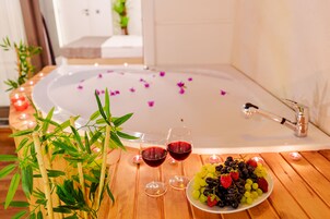 Una mesa de madera con un cuenco de fruta, dos copas de vino tinto y una bañera llena de pétalos de rosa.