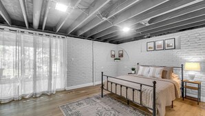 Un dormitorio con estructura de cama metálica, suelo de madera, pared de ladrillo blanco y techo con vigas a la vista.