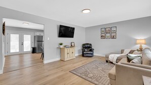 Una sala moderna con un sofá, una mesa de centro y un televisor montado en la pared. Se aprecia una cocina al fondo con un refrigerador y armarios.