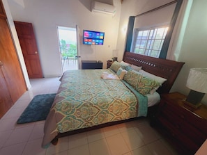 Un dormitorio con una cama grande, un televisor montado en la pared, una ventana con cortinas y una puerta que da a un espacio exterior.