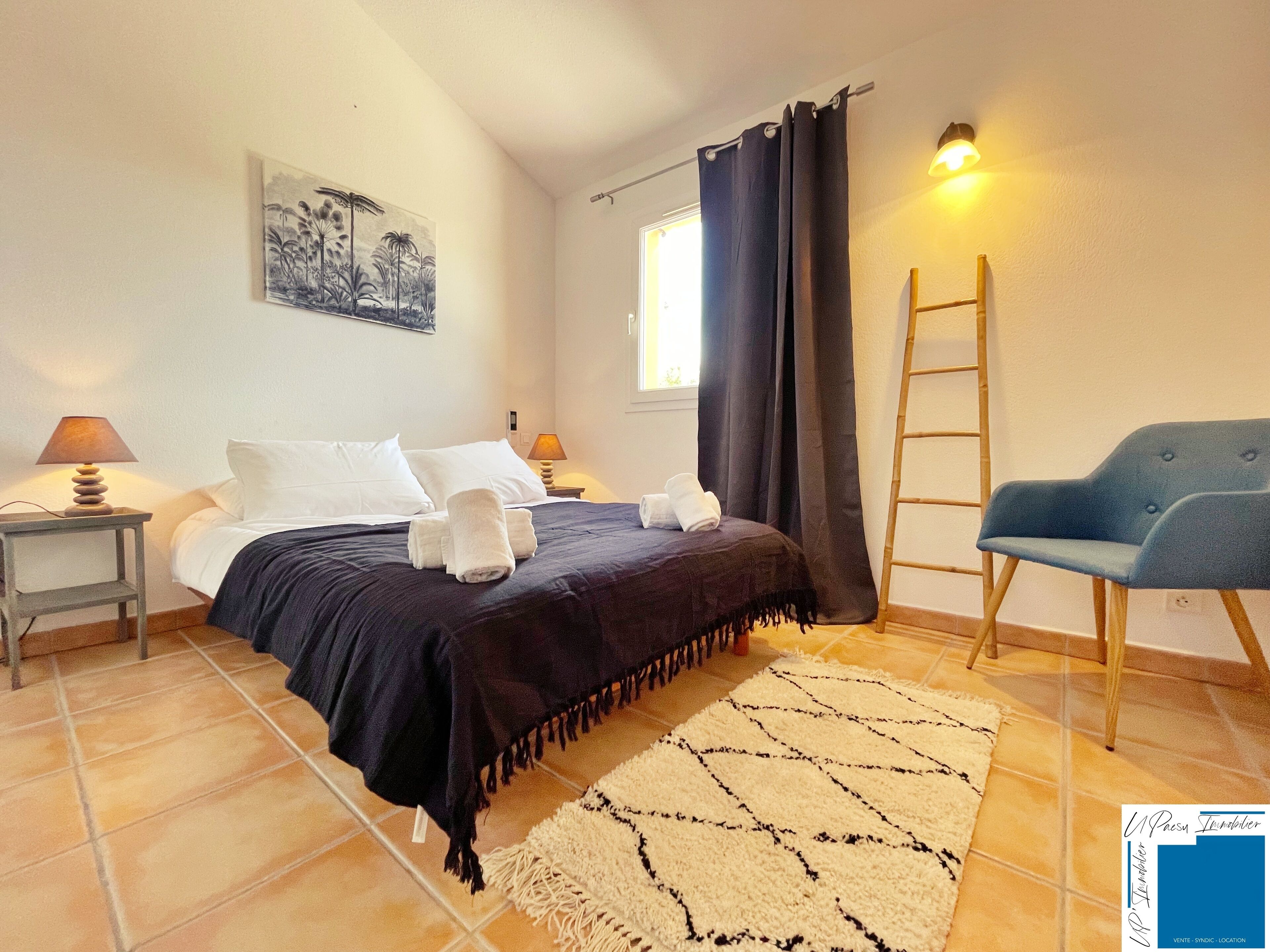 Quarto com cama, duas mesas de cabeceira com luminárias, uma cadeira, janela com cortinas, escada e tapete.