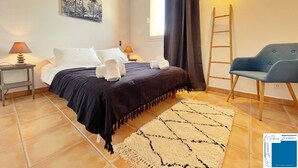 Ein Schlafzimmer mit einem Bett, zwei Nachttischen mit Lampen, einem Sessel, einem Fenster mit Vorhängen, einer Hockerleiter und einem Teppich.