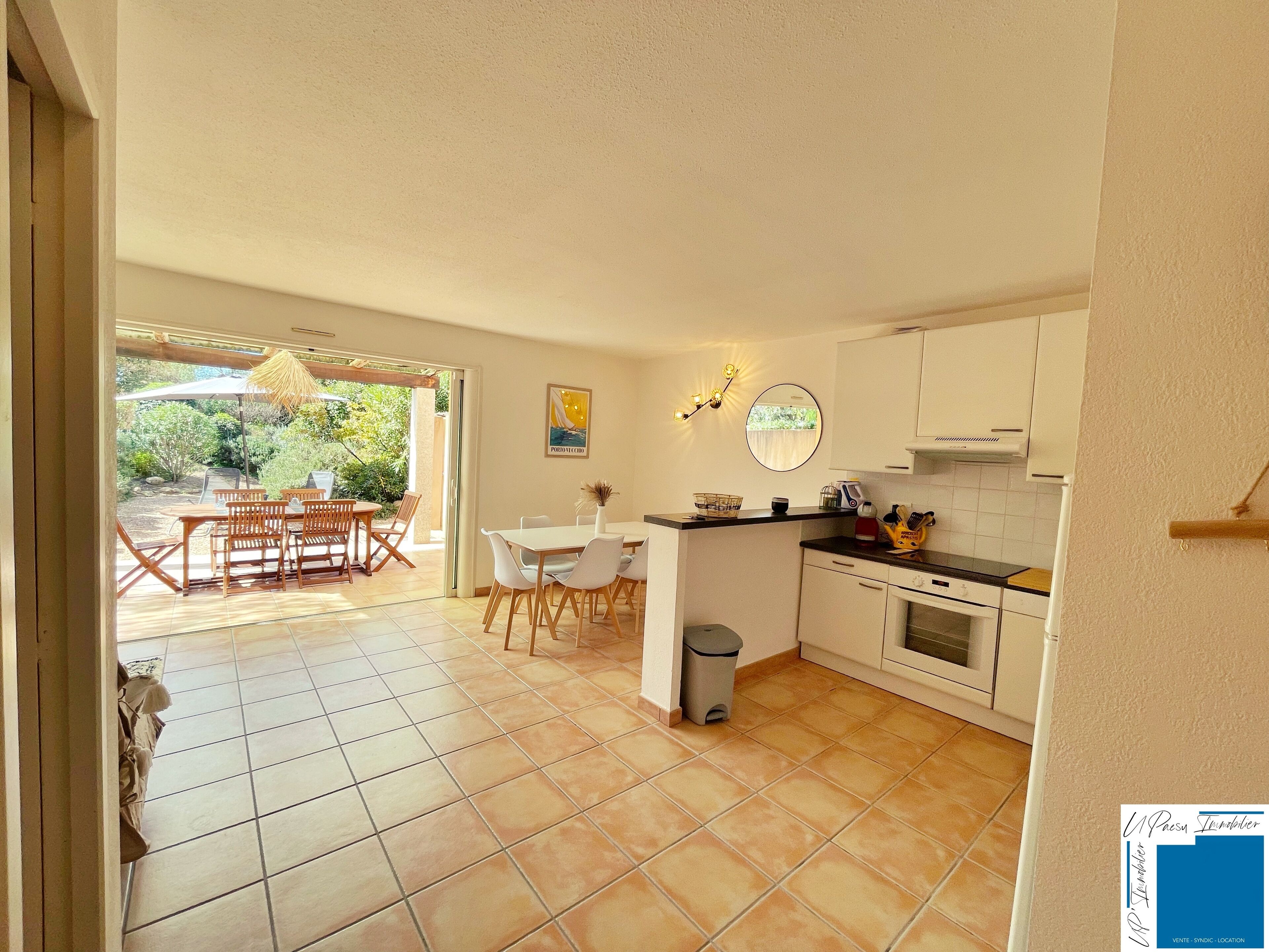 A compact kitchen with white cabinets, a dining area with a table and chairs, and a view of the garden through the open door.