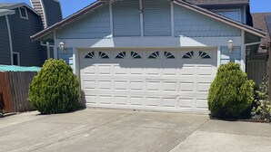 Un garage attenant à une maison, avec une porte blanche, flanqué de deux petits arbres, et une allée bien dégagée menant à la maison.