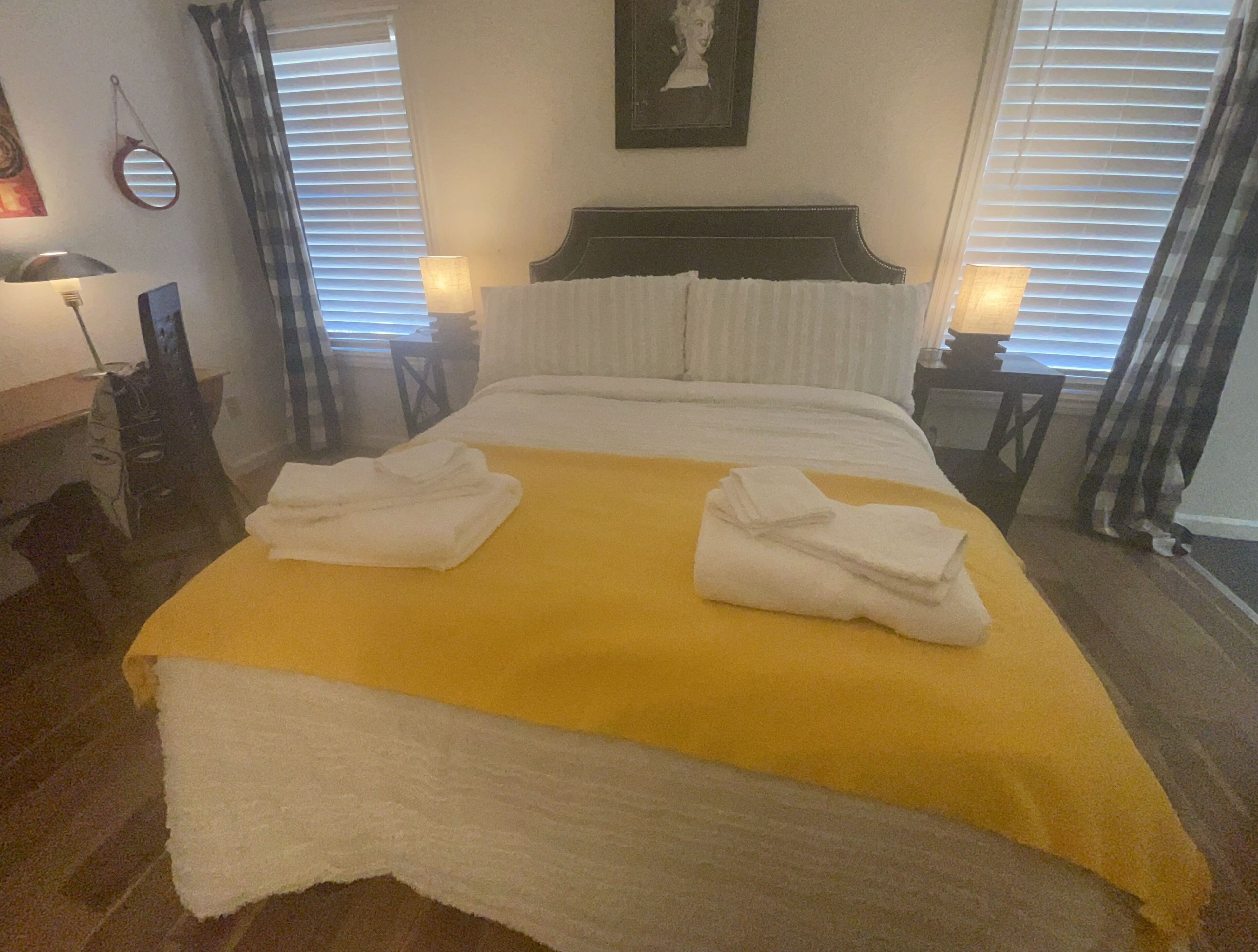 A neatly made bed with a yellow bedspread and two folded white towels, a framed portrait on the wall, and two bedside lamps.