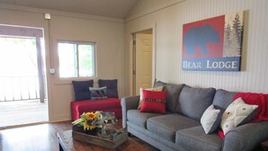 A living room with a grey sofa, a wooden coffee table, and a red ottoman. There is a window with blinds, a door, and a wall-mounted sign that reads "Bear Lodge". - Arrowhead Lake Camp - Black Bear Lodge Lake View (Lupton)