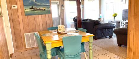 A dining area with a wooden table and chairs, a painting on the wall, and a view into the living room.