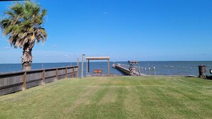 Un muelle de madera que se adentra en un mar tranquilo, una palmera a la izquierda y una zona de estar cubierta en el centro.