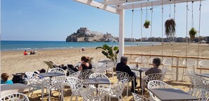 Un café situé en bord de mer, avec une terrasse offrant une vue sur l’océan et un château historique en arrière-plan.