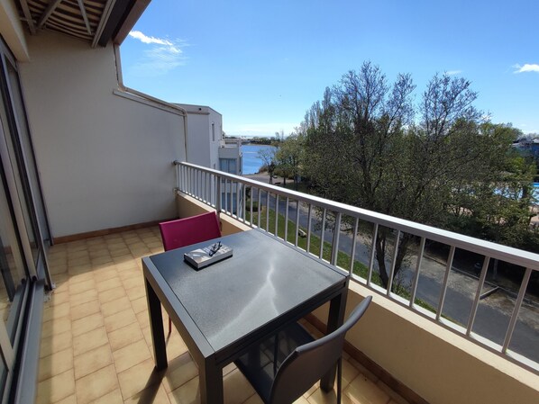 A balcony with a table and chairs, a view of a road and trees, and a building in the background.