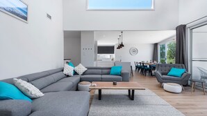 A modern living room with a grey sectional sofa, a wooden coffee table, and a dining area with a large window. - SeaSalt By The Sea Narrawallee (Narrawallee)