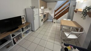 Een compacte keuken met een koelkast, magnetron en spoelbak. Er is een houten tafel met stoelen, een klein plankje met spullen, en een televisie op een standaard.