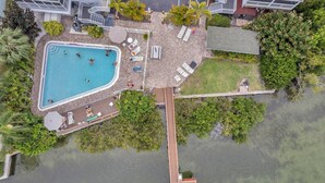 An aerial view of a resort with a swimming pool, lounge chairs, and a wooden deck extending over water.