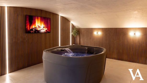 Un espace spa moderne doté d’un jacuzzi, de lambris en bois, d’un téléviseur diffusant une image de cheminée et de luminaires fixés au mur.