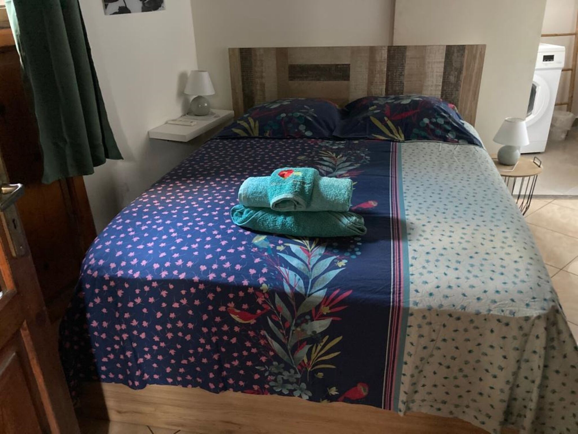 Een net opgemaakt bed met bloemenbedrukking, een opgevouwen handdoek en een nachtkastje met een lamp.