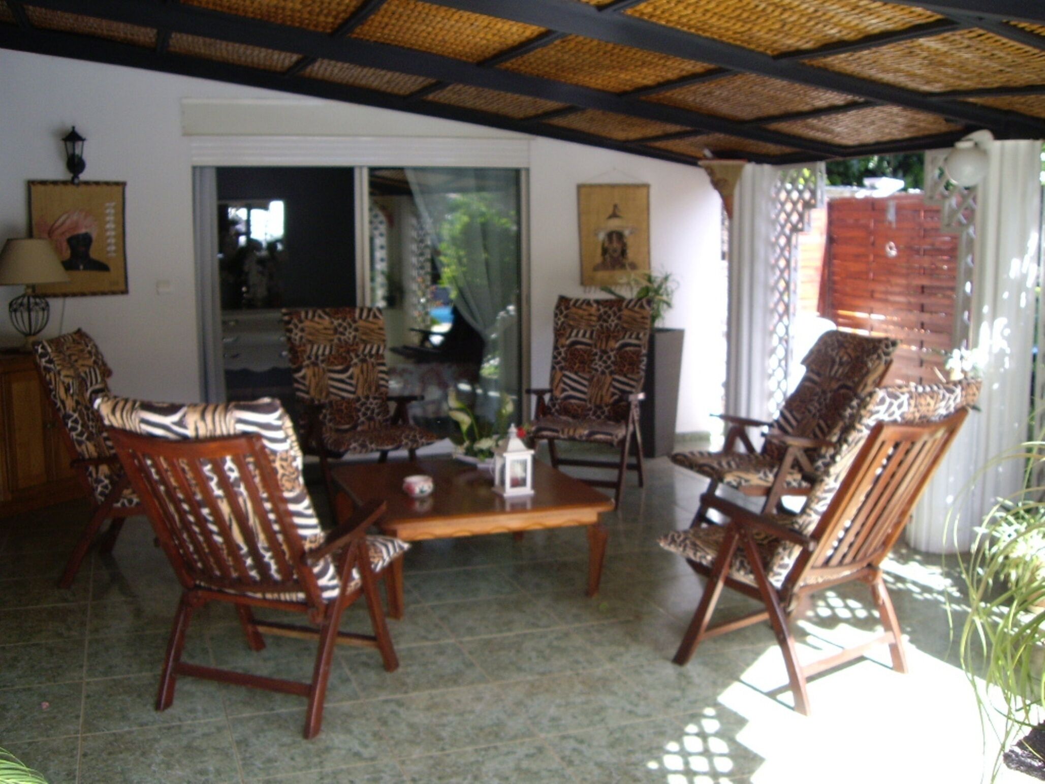 A room with a thatched ceiling, wooden chairs with patterned cushions, a wooden table, and a view through a glass door.
