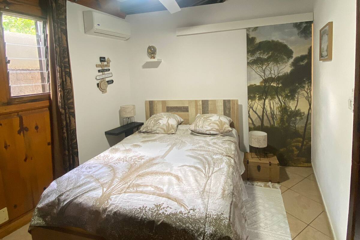 Un dormitorio con cama, ventilador de techo, mesita de noche, lámpara, un cuadro y un armario de madera.