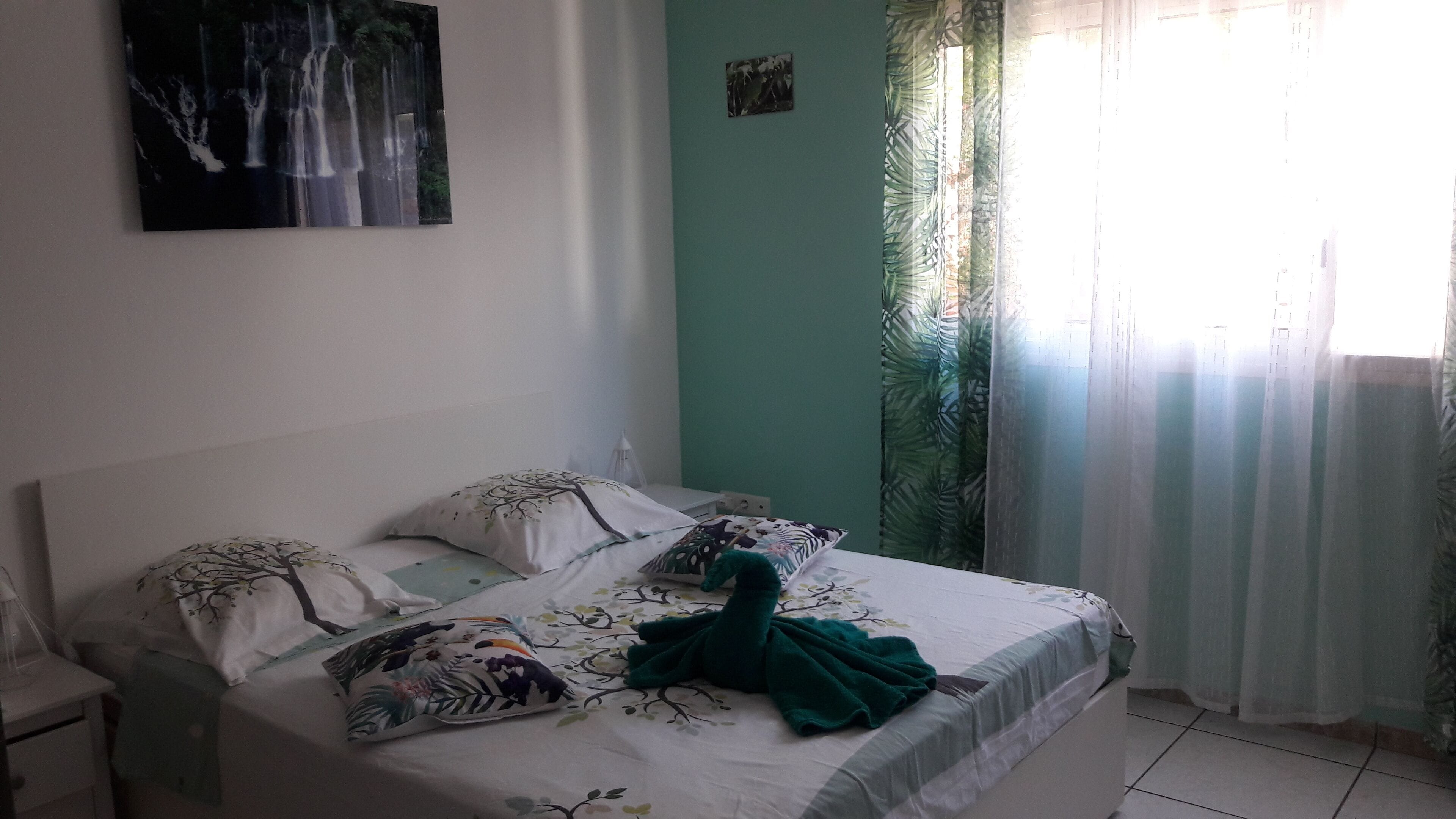 Ein Schlafzimmer mit Bett, Kissen, einem Nachttisch, einem Wandbild und einem Fenster mit Vorhängen.