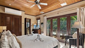 Ein Hotelzimmer mit einem großen Bett, einem Deckenventilator, einer Klimaanlage, einem Fernseher, einem Schreibtisch mit Stuhl und einem Balkon mit Blick auf Palmen.