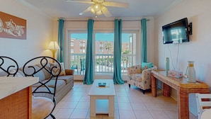 A well-lit living room with a ceiling fan, a TV mounted on the wall, and a view of the outside balcony through large windows. - Barefootin'-Victorian-Gulf Front-Sleeps 6 (Galveston)