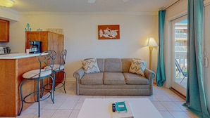 A living room with a sofa, a coffee table, a dining area with a chair, a kitchen area with a coffee maker, and a balcony with curtains. - Barefootin'-Victorian-Gulf Front-Sleeps 6 (Galveston)