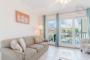 Un salon avec un canapé beige, une table basse et une vue sur une piscine et des immeubles, visible à travers de grandes fenêtres.