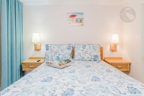 A bed with a floral pattern, two bedside lamps, a small bedside table with a remote control, and a wall-mounted picture.