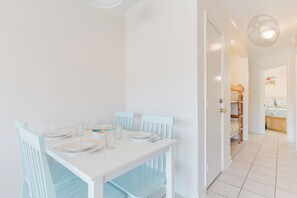 Un comedor con una mesa y sillas blancas, una puerta que da a un dormitorio y un pasillo con una cama y un ventanal.