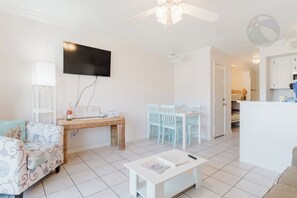 A modern living room with a flat-screen TV, a dining area with white chairs, a white coffee table, and a kitchen area with white cabinets. - Barefootin'-Victorian-Gulf Front-Sleeps 6 (Galveston)