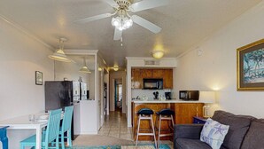 Een ruimtelijk ingerichte leefruimte met een kitchenette, een eethoek met tafel en stoelen, een bank en een plafondventilator.