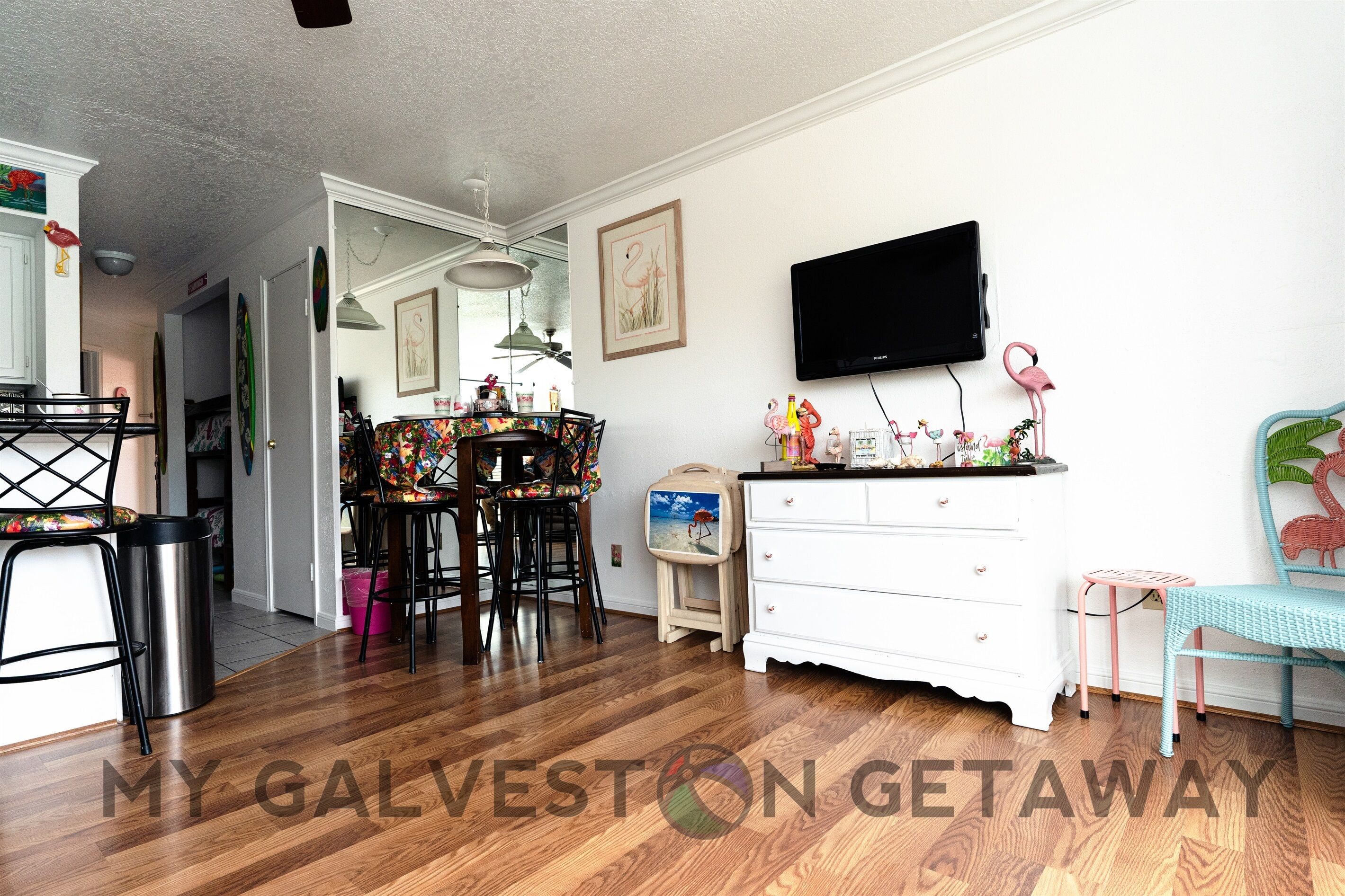 A dining area with a wooden table and chairs, a white dresser, and a television mounted on the wall.