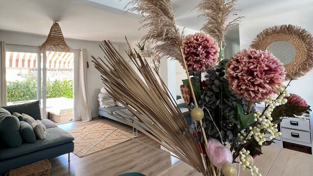 A modern living room with a wooden dining table, a sofa, and a vase with dried plants and flowers.