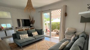 A modern living room with a grey sofa, a television, and a kitchen area visible through an open sliding door.