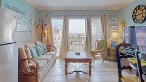 A cozy living room with a sofa, a wooden table, and a chair. There is a balcony with curtains and a view of the outside. - Makin' Memories @ Victorian (GVR05004) (Galveston)